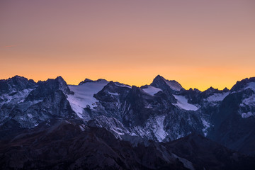 Fototapeta na wymiar Colorful sky at dusk beyond the glaciers on the majestic peaks of the Massif des Ecrins (4101 m), France. Telephoto view from distant at high altitude. Clear orange sky.