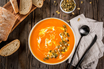 Traditional fall and winter dishes, hot and spicy pumpkin  soup with pumpkin seeds, cream and freshly baked baguette, on old rustic wooden table, copy space top view
