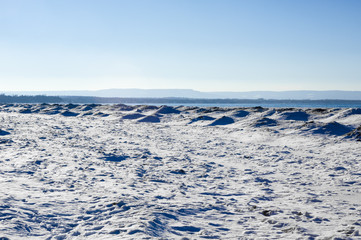 Frozen ice, snow, and sand dunes on beach with hills in distance.