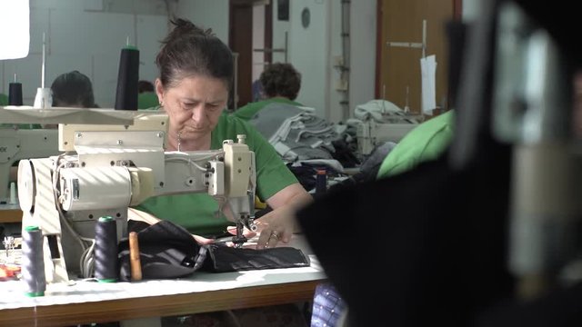 Close up of middle aged Caucasian woman during sits behind sewing machine and stitching factory textile, group of woman working in the blurred background, shallow depth of field, interior scene