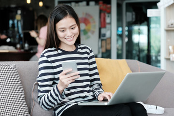 Beautiful asian girl using laptop computer and smart phone while sitting on sofa with smiling face emotion, people and technology concept, lifestyle