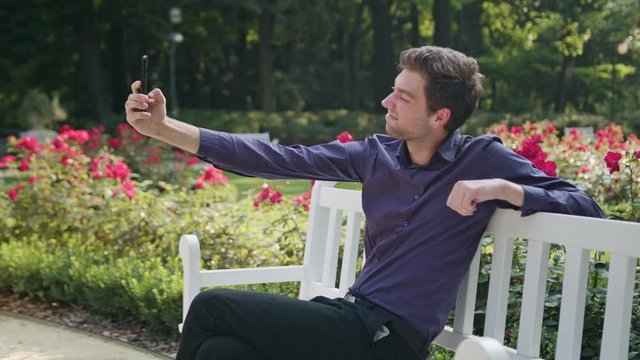 A young man sitting on a white bench in the park and using a phone to make a selfie. Medium shot
