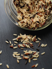 Close up uncooked mixed rice grains falling out of canning jar on black, wooden background containing white long grain rice, whole brown rice, black rice and black wild rice