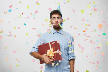 Unhappy manager with stubble wearing holiday cap posing in studio with box of birthday gift in...