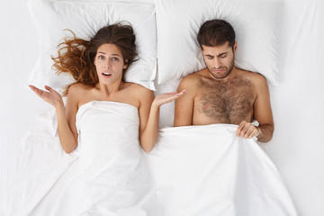 Displeased indignant Caucasian woman shrugging shoulders with open palms after her husband couldn't perform sexually again. Frustrated male having erectile dysfunction, lying in bed with his wife