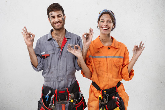 Picture of young female and male workers of maintenance service wearing working clothes and waist bags with tools, smiling, making mudra gesture, trying to stay clam and positive about their hard work