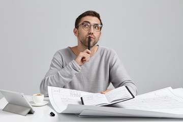 Naklejka premium Waiting for inspiration. Horizontal shot of handsome talented European designer making notes in copybook, looking sideways, holding black pen at his lips, writing down creative ideas and solutions
