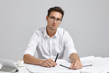 Confident handsome male architect, wears formal white shirt, round spectacles, holds pen, generates new ideas, writes notes in notebook, sits at work place, surrounded with blueprints, gadget, coffee