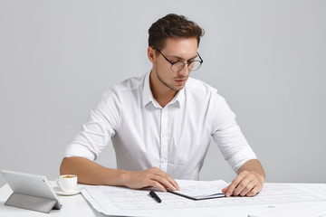 Serious businessman looks at diary, decides when to arrange meeting, sits in office, work with modern tablet, drinks cappuccino, isolated over grey background. People, occupation, business concept