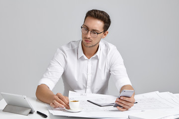 Candid shot of concentrated talented engineer wearing white formal shirt and round glasses sitting at office desk, focused on work, checking blueprints, using electronic devices and drinking espresso