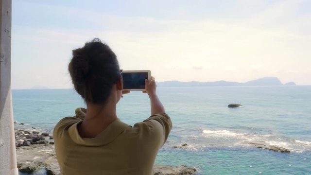 Young Woman in a wooden veranda taking pictures of the sea with her smartphone from a cliff on a desert Island in Italy