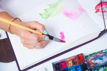 A close-up as an artist with blue nails draws pink flowers with a wooden brush and watercolors in the aluoma for drawing, on the table lie a palette for drawing