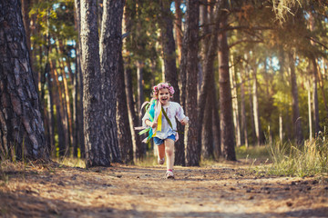 A little girl in a wreath with ribbons is running happily through the summer forest.