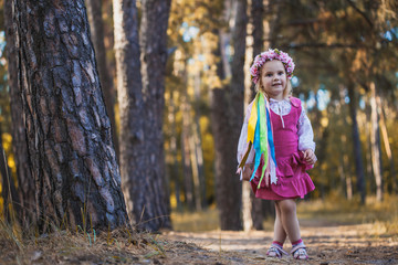 A little girl in a wreath with ribbons is running happily through the summer forest.