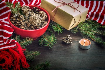 Christmas decoration - red bowl full of fir-cones, gift box wrapped in kraft paper, pine branches, candle and red and white striped winter scarf. Christmas theme. Dark wooden background.