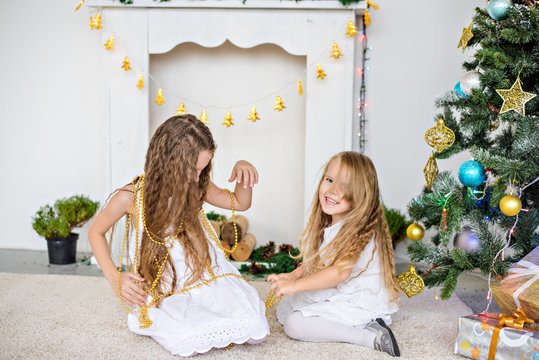 Two little blonde girls in white dresses are playing by the fireplace and Christmas tree with boxes. Sisters have fun, hug, laugh, unfold gifts from Santa Claus. New year card. Copy space. Friendship