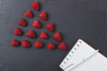 Red raspberries, tape measure, notebook and a pen on the black slate stone background