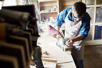Portrait of skilled carpenter polishing piece of wood in workshop standing against window in sunlight and  making furniture