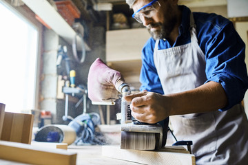 Portrait of mature bearded carpenter making furniture in workshop, polishing piece of wood standing against window in sunlight, focus on sanding machine