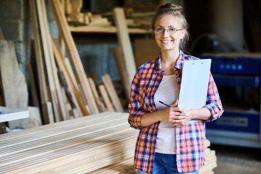 Portrait of happy young woman wearing plaid shirt posing in woodworking shop smiling looking at camera and holding clipboard, copy space