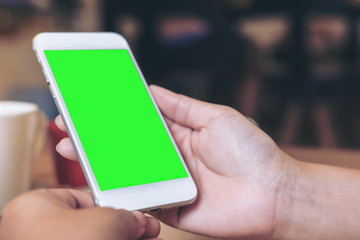 Mockup image of hands holding white mobile phone with blank green screen with coffee cups on wooden table in restaurant