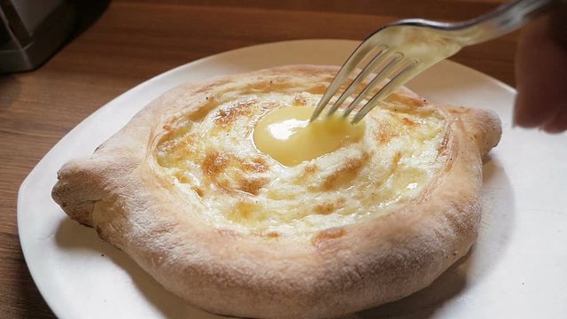 Female hand mixing ingredients of adjarian khachapuri with fork. Khachapuri is open bread stuffed with cheese and egg yolk, traditional dish of Georgian cuisine. Close up