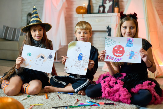 Positive cute friends in Halloween costumes sitting on carpet with splattering markers and showing creative pictures with Halloween characters to camera at home party