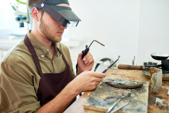 Side view portrait of young man holding small gas torch and welding tools while working with metal against shabby wooden workstation
