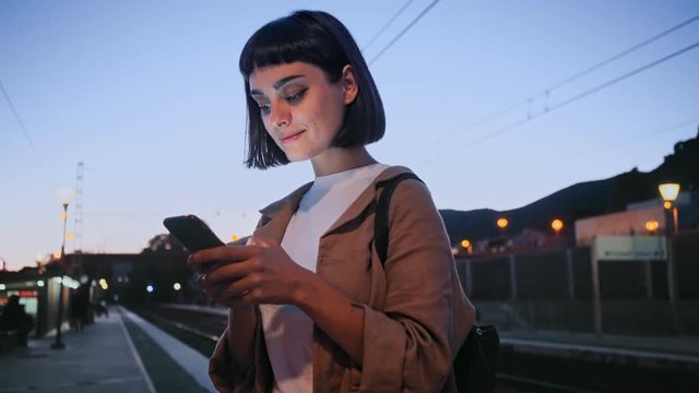 Beautiful and attractive young woman stands on station platform in night time, uses smartphone, to browse internet or scroll through news feed, fast two floor train express passes by