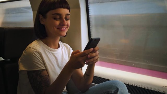 Attractive positive woman in white tshirt and jeans, chats on messaging application or plays online game, she sits in fast moving high speed train during morning commute