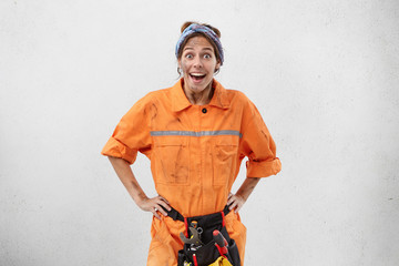 Joyful female plumber wears uniform, being glad after successful repair of clogged stink, wonders her professionalism, looks confidently, keeps hands on waist, waits for new orders from clients