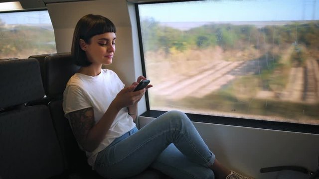 Cute young millennial hipster woman or teenager with cool arm tattoos sits in fast train, next to window, landscapes pass by, she uses smartphone and texts smiling and chuckling