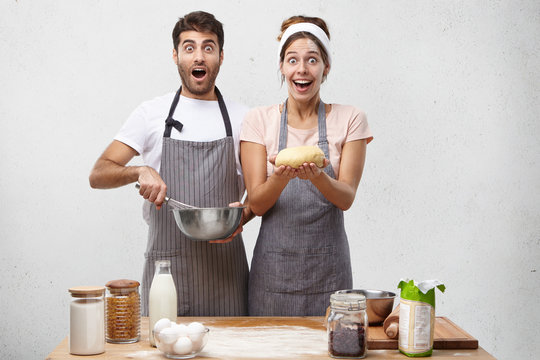 Astonished male anf female cookers look with jaw dropped out and widely opened eyes, make pastry for baking delisious dessert, being shocked to see guests at threshold, haven`t prepare dinner yet