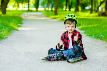 Boy in a protective helmet and protective pads for roller skating sitting on the road and showing thumbs up