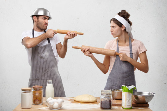 Two female and male competitors struggle for winning culinary contest, target at each other with rolling pins, have irritated and furious expressions, use kitchen supplies as weapon for defense