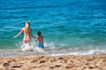Caucasian mom and son running into the sea water. Back view.