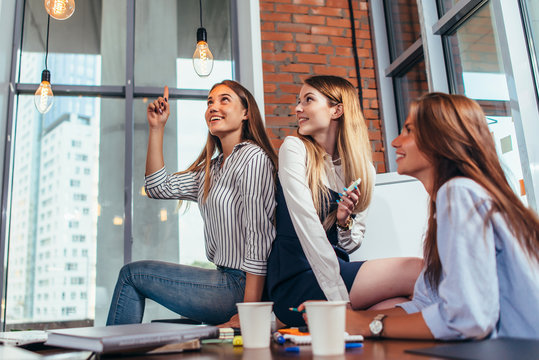 Happy Female Teenagers Sitting On Table Looking And Pointing At Stylish Light Bulbs And Relaxing During A Recess Between The Lessons In Classroom