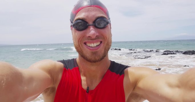 Triathlon swimming man - male triathlete swimmer selfie video wearing swim goggles and cap after ocean swim training session. Fit man in professional triathlon suit training for ironman.