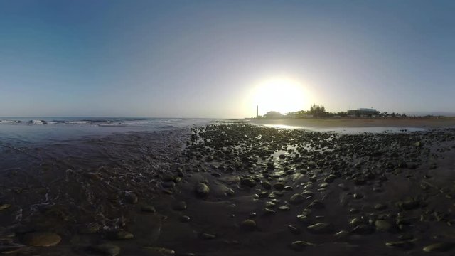 Ocean coast with waves washing rocks. View to the resort with Maspalomas Lighthouse against setting sun, Gran Canaria