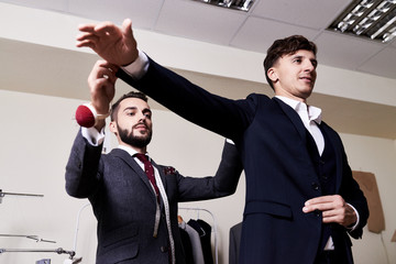 Low angle view of stylish young tailor fitting custom-made suit to handsome client, interior of...