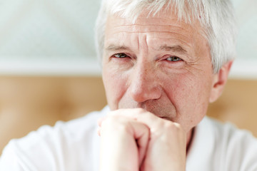Grey-haired man with wrinkled face keeping his hands by mouth while staring at camera