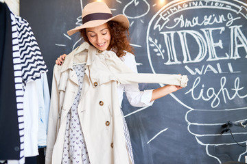 Portrait of happy young woman trying on new clothes in store, holding autumn trench coat and hat