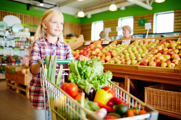 Cute little girl pushing shopping cart with fresh vegetables in supermarket