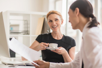 Portrait of joyful blonde entrepreneur holding cup of coffee in hands while discussing details of mutually beneficial cooperation with business partner at modern boardroom