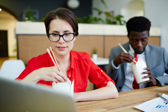 Multi-ethnic group of busy colleagues eating instant noodles with chopsticks while working at modern open plan office
