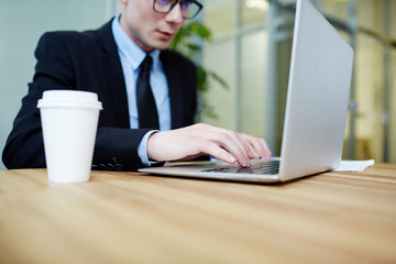 Close-up shot of confident young manager wearing suit sitting in front of laptop and writing response email to client, interior of spacious open plan office on background