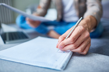 Close-up shot of unrecognizable entrepreneur sitting on cozy bed and taking necessary notes while preparing for important negotiations with business partner, blurred background