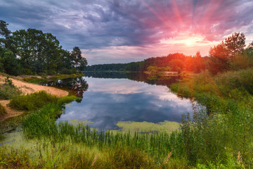 Fototapeta na wymiar Beautiful landscape of the lake at colorful sunset. Dramatic sky in orange and purple colors. Panoramic view of the backwater and green coast with trees reflecting in the water.