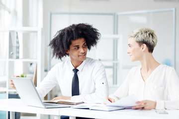 Obraz premium Cheerful young colleagues looking at each other with warm smiles while having project discussion at spacious open plan office