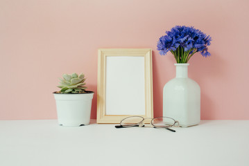 Home office desk with photo frame mock up, bouquet of cornflowers in front of pale pink pastel background. Social media concept